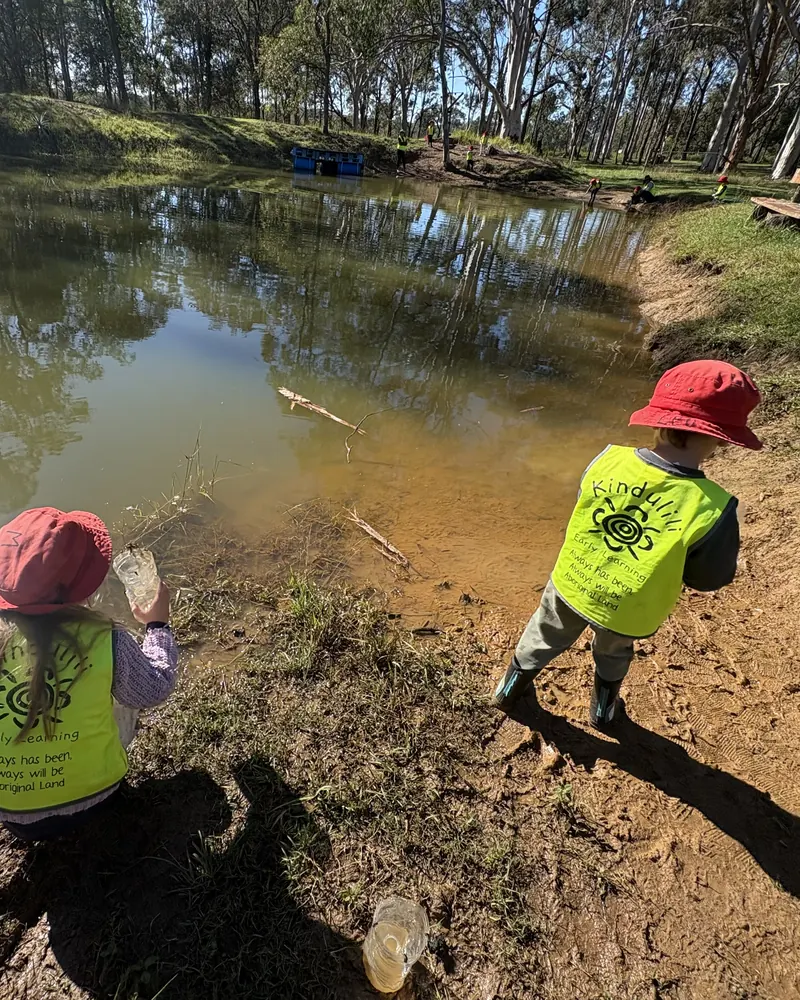 Two children in red hats and hi-vis vests exploring the edge of a bush dam